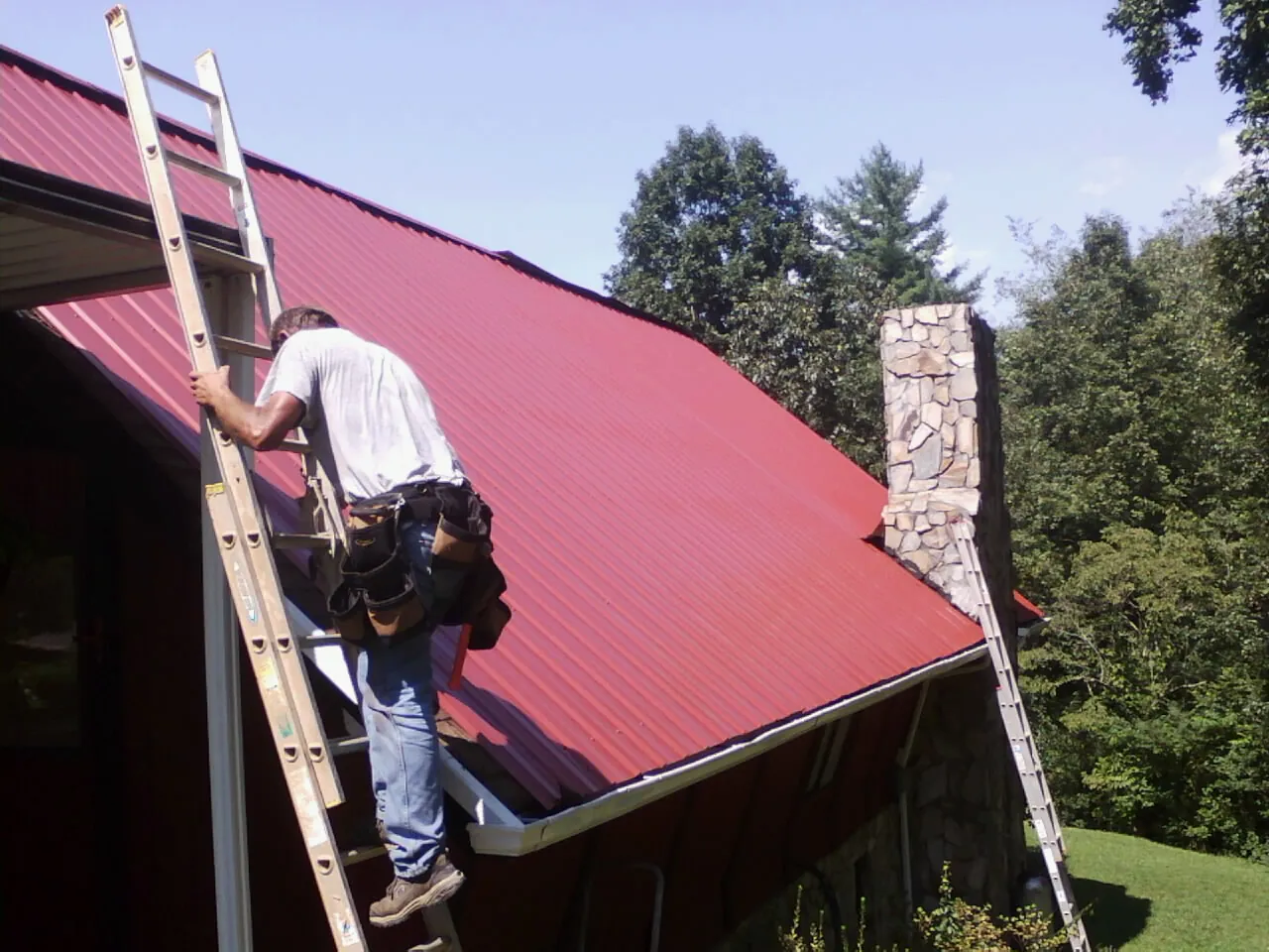 Roofer on a ladder beside a red metal roof and stone chimney — Soffit & Fascia Repair by Roofing Brothers in Gobles